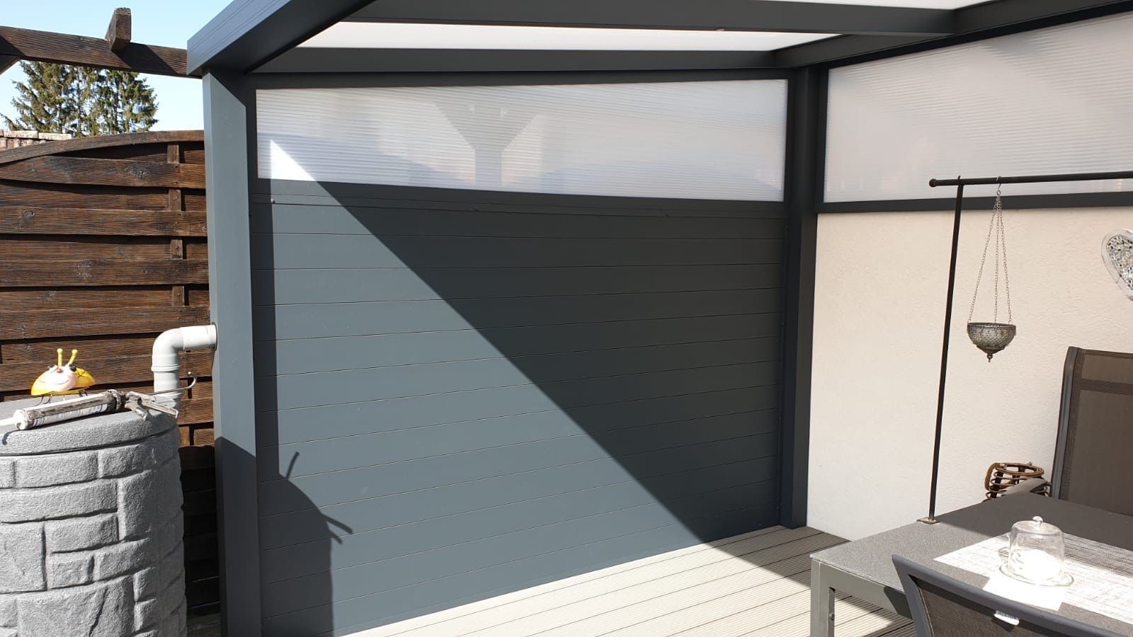 Blick auf die Terrasse mit einer dunkelgrauen Sichtschutzwand, deren oberer Teil unter einer modernen Pergola-Konstruktion lichtdurchlässig ist.