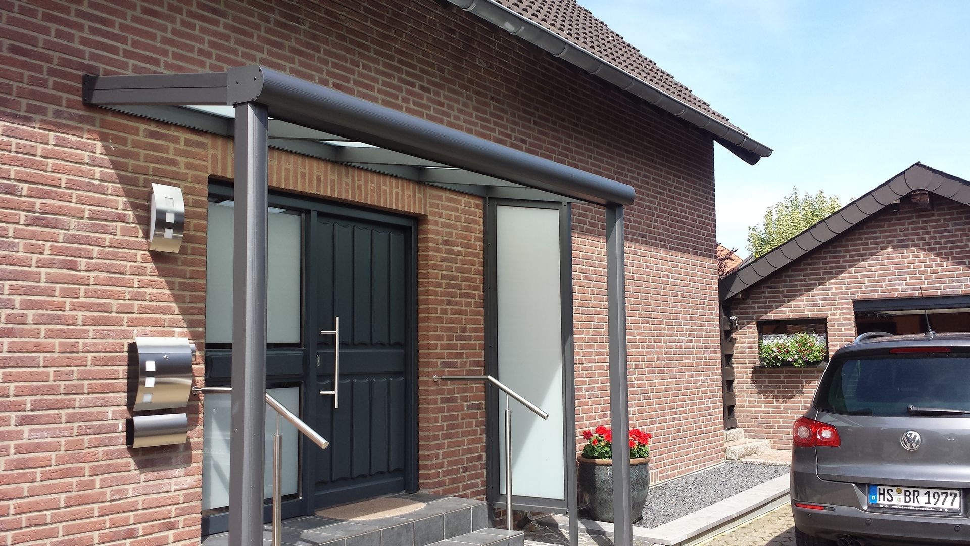 Ein dunkelgrauer Hauseingang mit Glasvordach, Backsteinwänden, einem Handlauf und einem seitlich geparkten Auto.