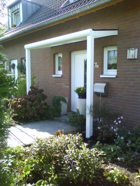 Ein gemauerter Hauseingang mit einem weißen Vordach, das von zwei weißen Säulen über einem Betonweg und einem Garten getragen wird.