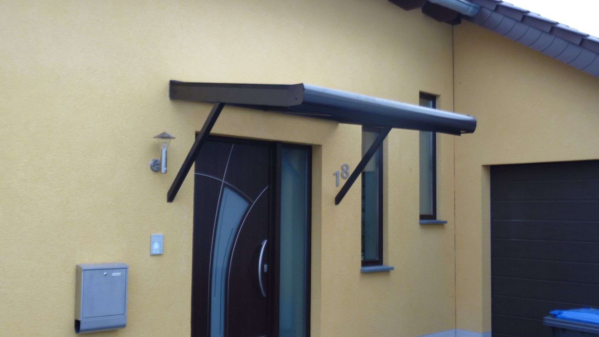 Ein gelber Hauseingang mit einer modernen dunkelbraunen Tür, einem rechteckigen Metallvordach und einem an der Wand montierten Briefkasten.