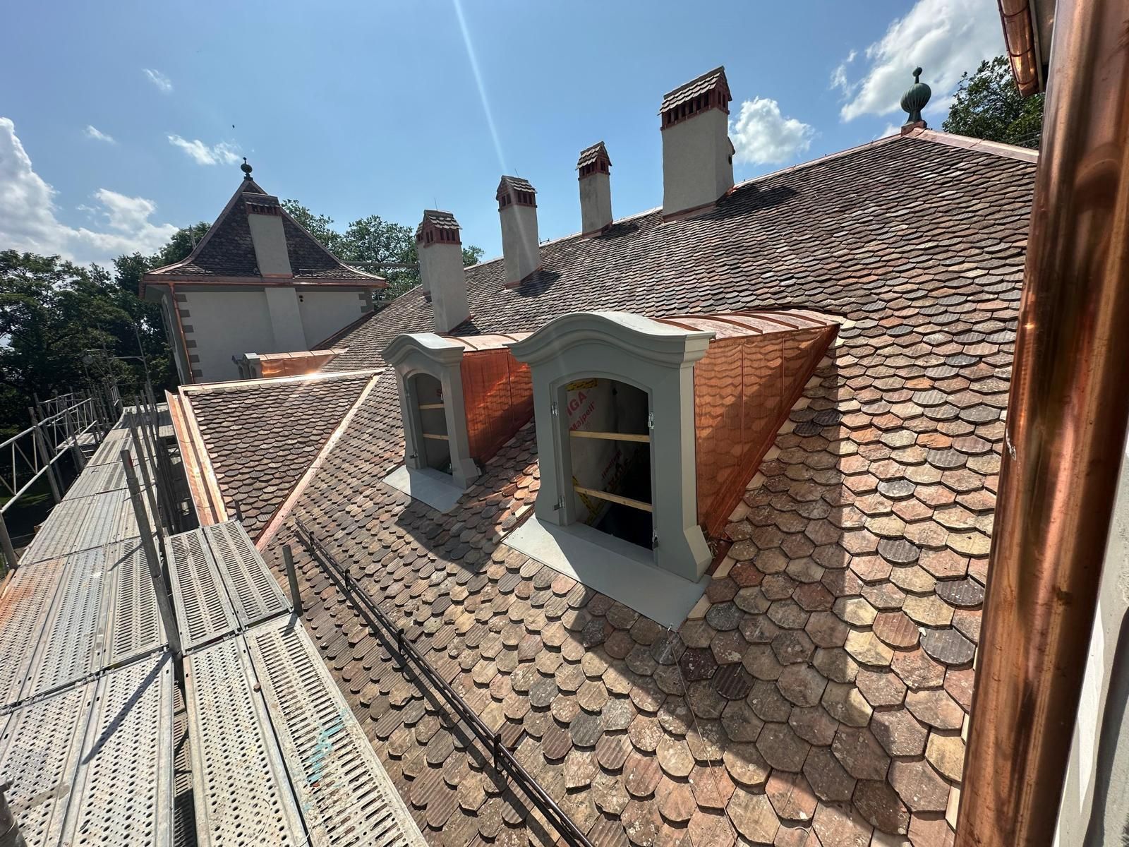 Toiture de tuiles avec lucarnes et cheminées sous un ciel bleu, à côté d'un bâtiment en pierre et d'échafaudages.