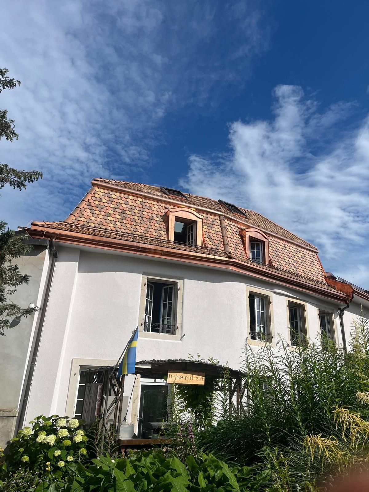 Maison blanche au toit de tuiles rouges et aux lucarnes, sous un ciel bleu, entourée de verdure.