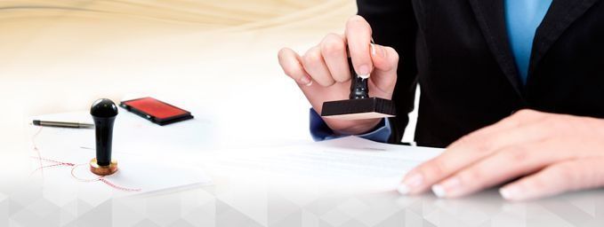 Persona sellando un documento con un sello de goma negro. Cerca hay otros materiales de oficina, como un bolígrafo y un tintero.