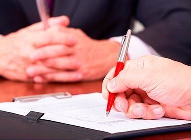 Persona firmando un documento con bolígrafo rojo, con otra persona al fondo. Primer plano.