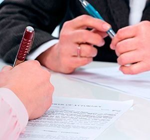 Dos personas en una mesa firman un documento. Una sostiene un bolígrafo rojo y la otra, uno azul.