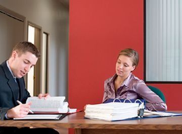Hombre de traje revisando documentos, mujer observando. Oficina con pared roja.