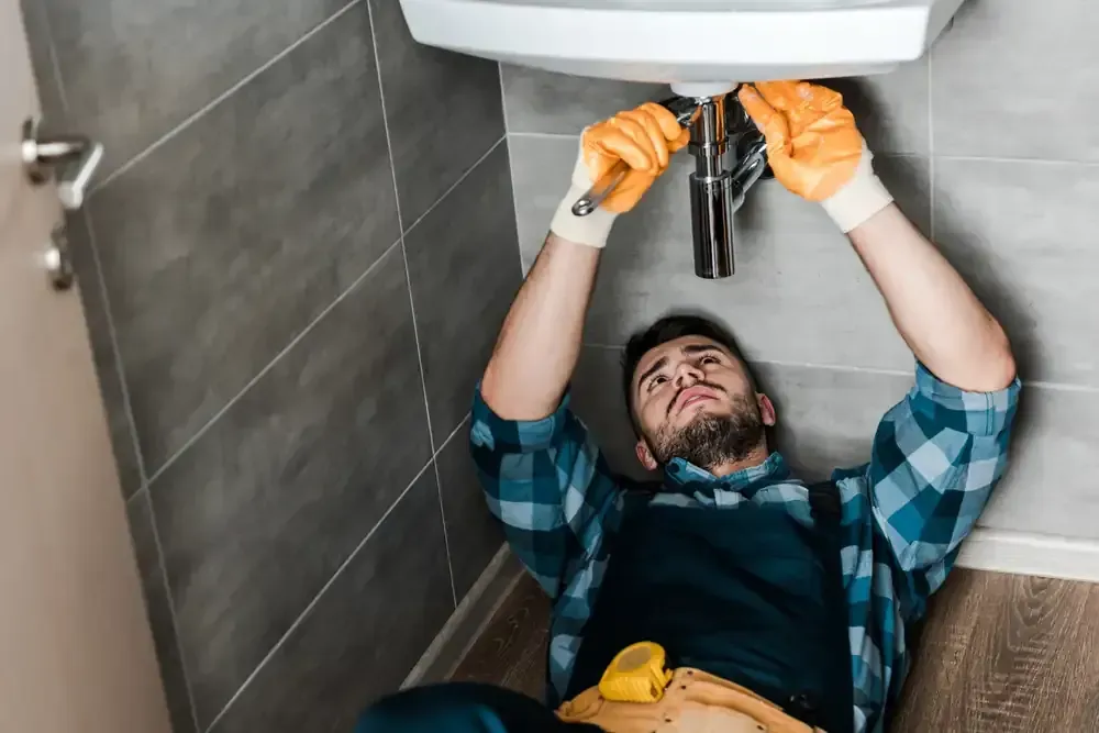 Fontanero con guantes trabajando bajo el lavabo del baño con una llave inglesa. El hombre lleva una camisa azul a cuadros.