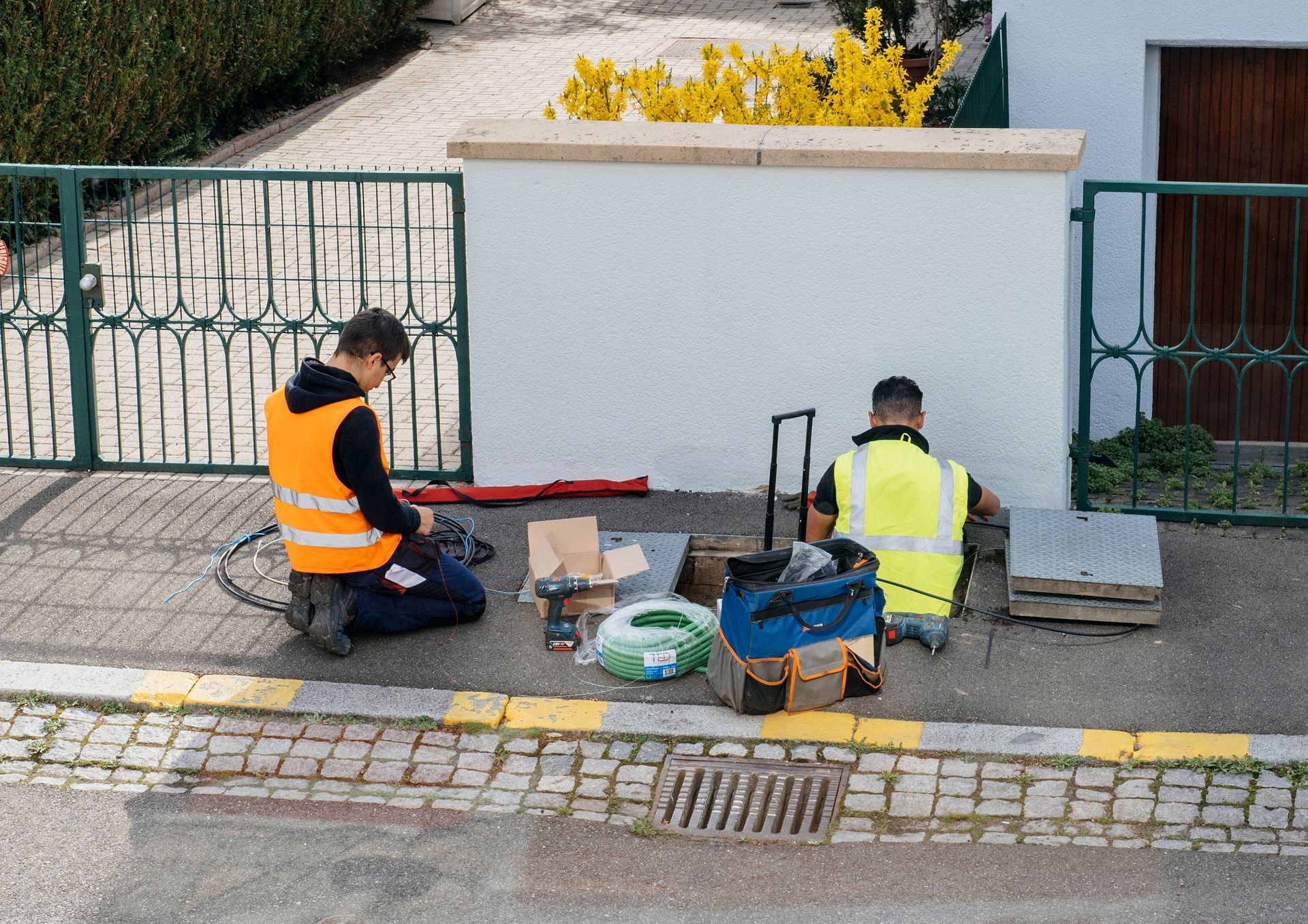 Dos trabajadores instalan equipos en una pared blanca cerca de una carretera. Llevan chalecos antibalas, están arrodillados
