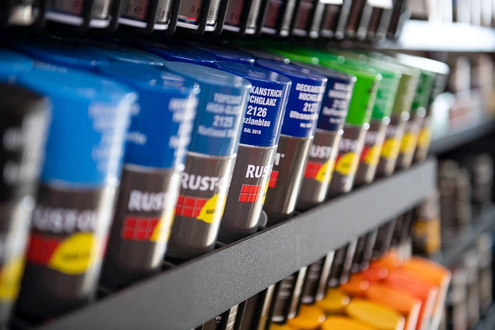 Reihen von Sprühdosen in verschiedenen Farben auf einem Ladenregal, darunter blau, grün und orange, mit dem Rust-Oleum-Logo.