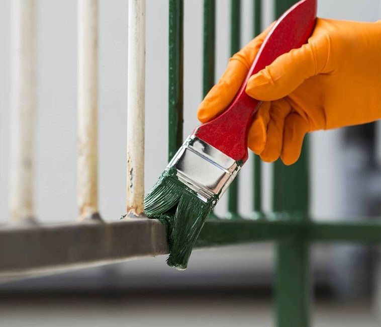 Eine Hand mit einem orangefarbenen Handschuh streicht mit einem Pinsel eine grüne Schicht Farbe auf ein weisses Metallgeländer.