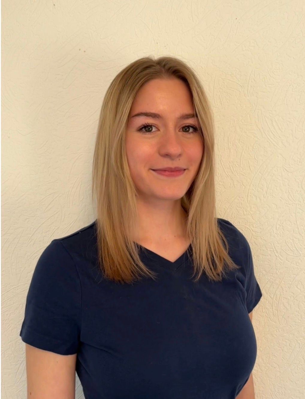 Eine Person mit glattem blonden Haar, die ein dunkelblaues T-Shirt trägt, steht vor einer hellen Wand.