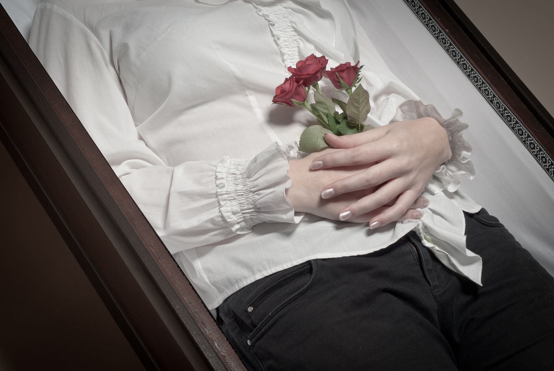 Une personne allongée dans un cercueil, tenant un petit bouquet de roses rouges, vêtue d'un chemisier et d'un pantalon.
