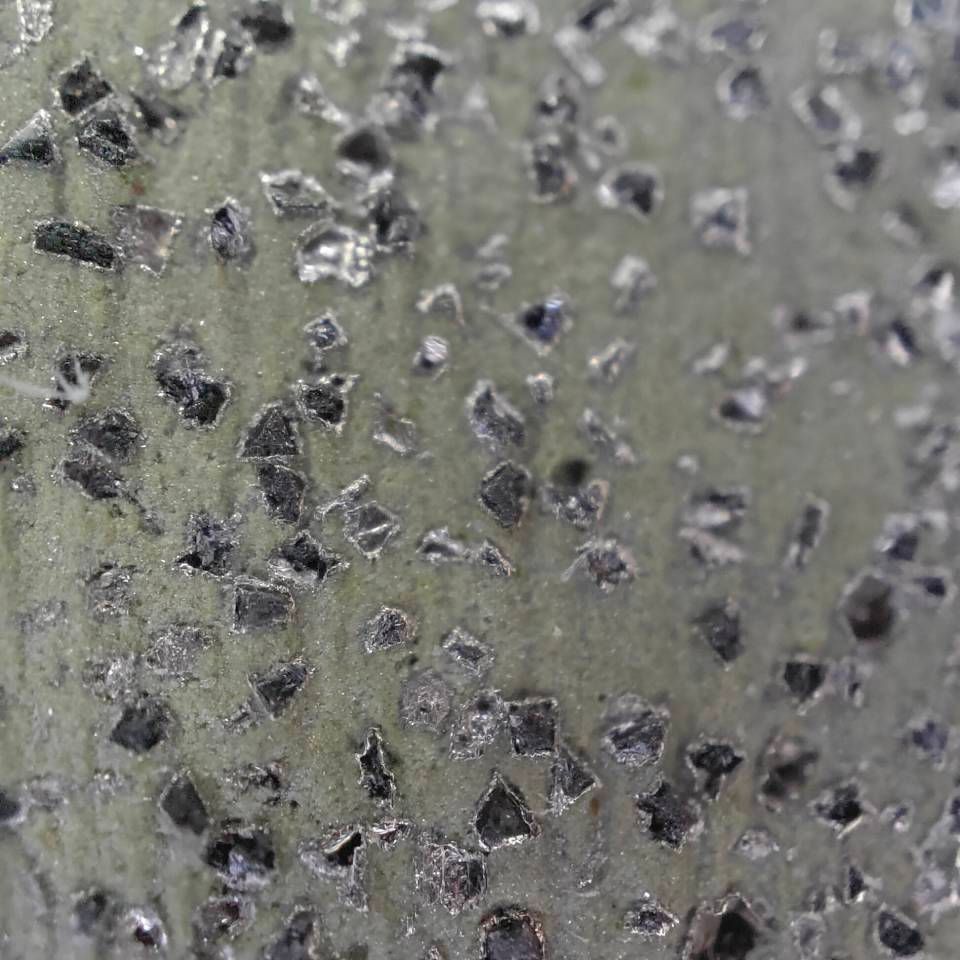 Nahaufnahme einer grünlich-grauen, strukturierten Oberfläche mit vereinzelten dunklen Flecken und Rissen
