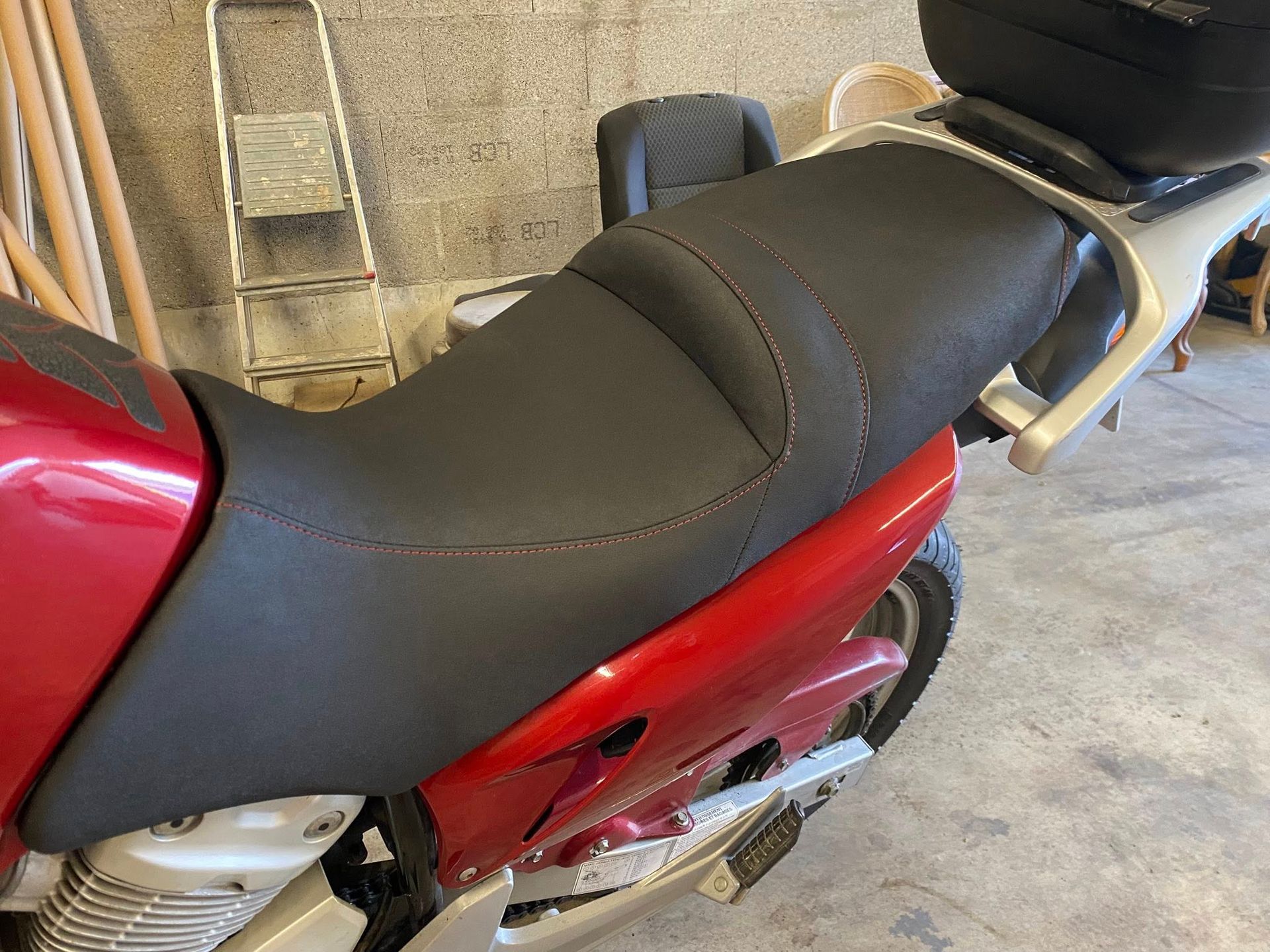 Selle de moto noire sur une moto rouge, avec un conteneur de rangement noir dans un garage.