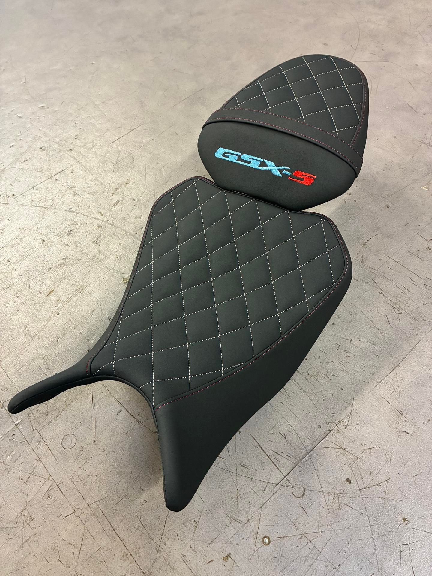 Selle de moto noire avec surpiqûres en losange, logo GSX-S et appuie-tête sur fond gris.