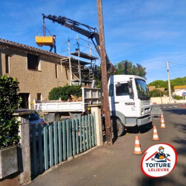 Un camion-grue soulève un conteneur de chantier sur le toit d'une maison équipée d'échafaudages, avec des cônes de signalisation au premier plan.