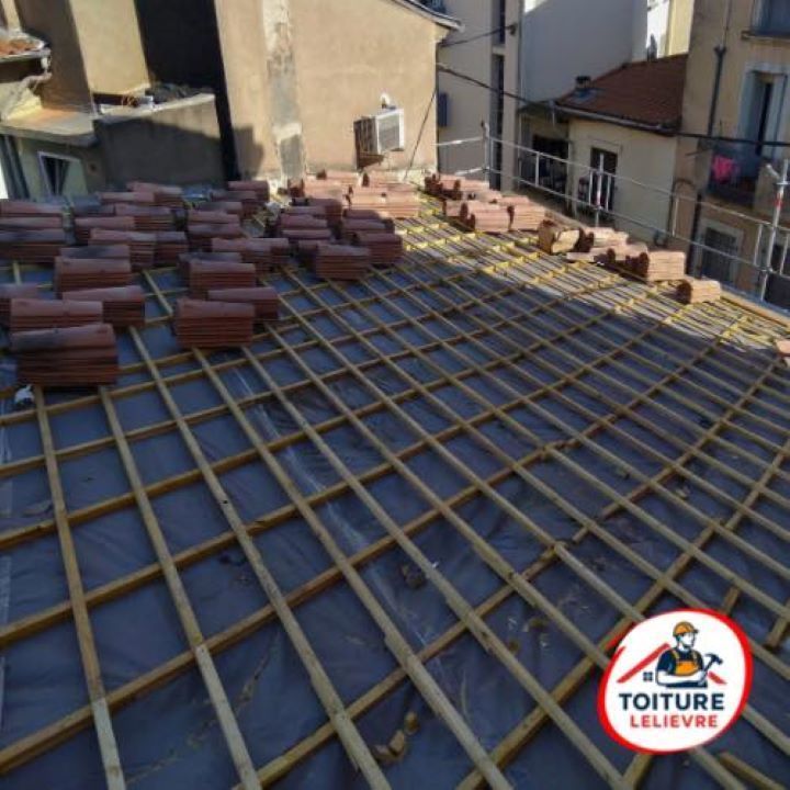 Une toiture en rénovation, présentant une grille de liteaux en bois installée sur une membrane protectrice sombre, avec des piles de tuiles.