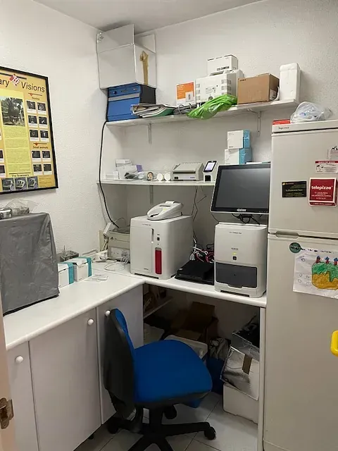 Una pequeña oficina con escritorio, equipo y espacio de almacenamiento. Paredes blancas, una silla azul y varios artículos en estanterías.