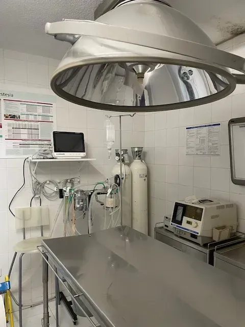Un quirófano con lámpara quirúrgica, mesa, monitores, tanques de oxígeno y una silla.