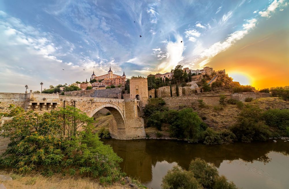 Puente medieval y edificios sobre un río, con un cielo al atardecer.