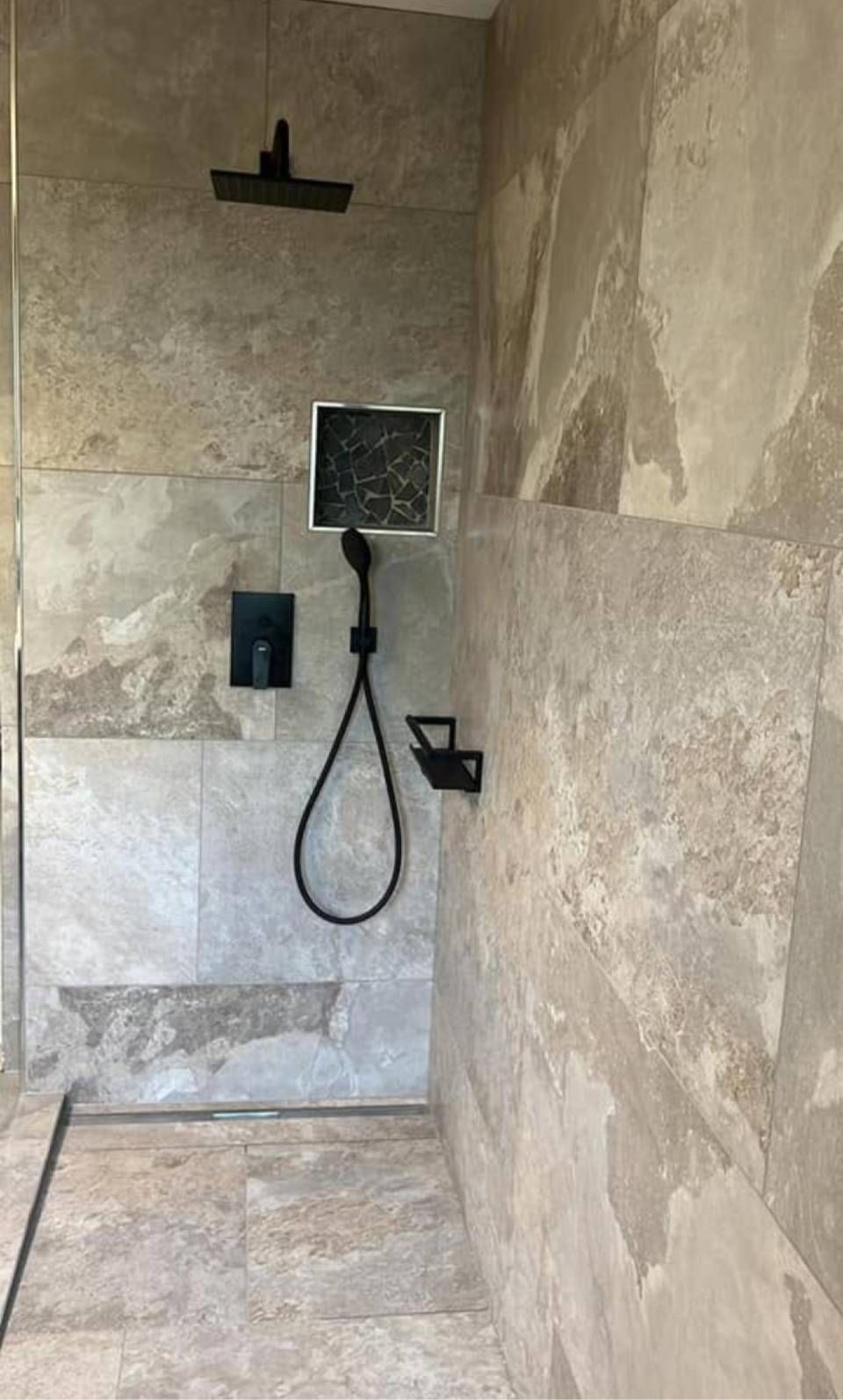 Begehbare Dusche mit beigen Fliesen, schwarzen Armaturen und einem an der Wand montierten Duschkopf.