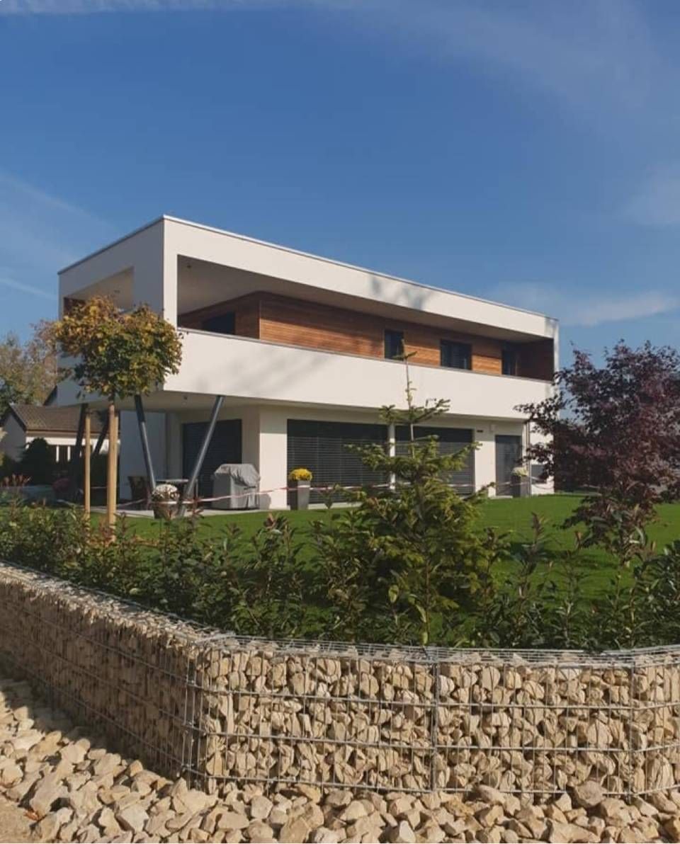 Modernes, weisses zweistöckiges Haus mit Balkon, hinter einer Steinmauer und einem grünen Garten unter blauem Himmel
