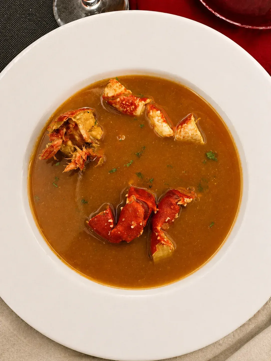 Un tazón blanco de sopa marrón cubierto con trozos de langosta roja y hierbas sobre una mesa.
