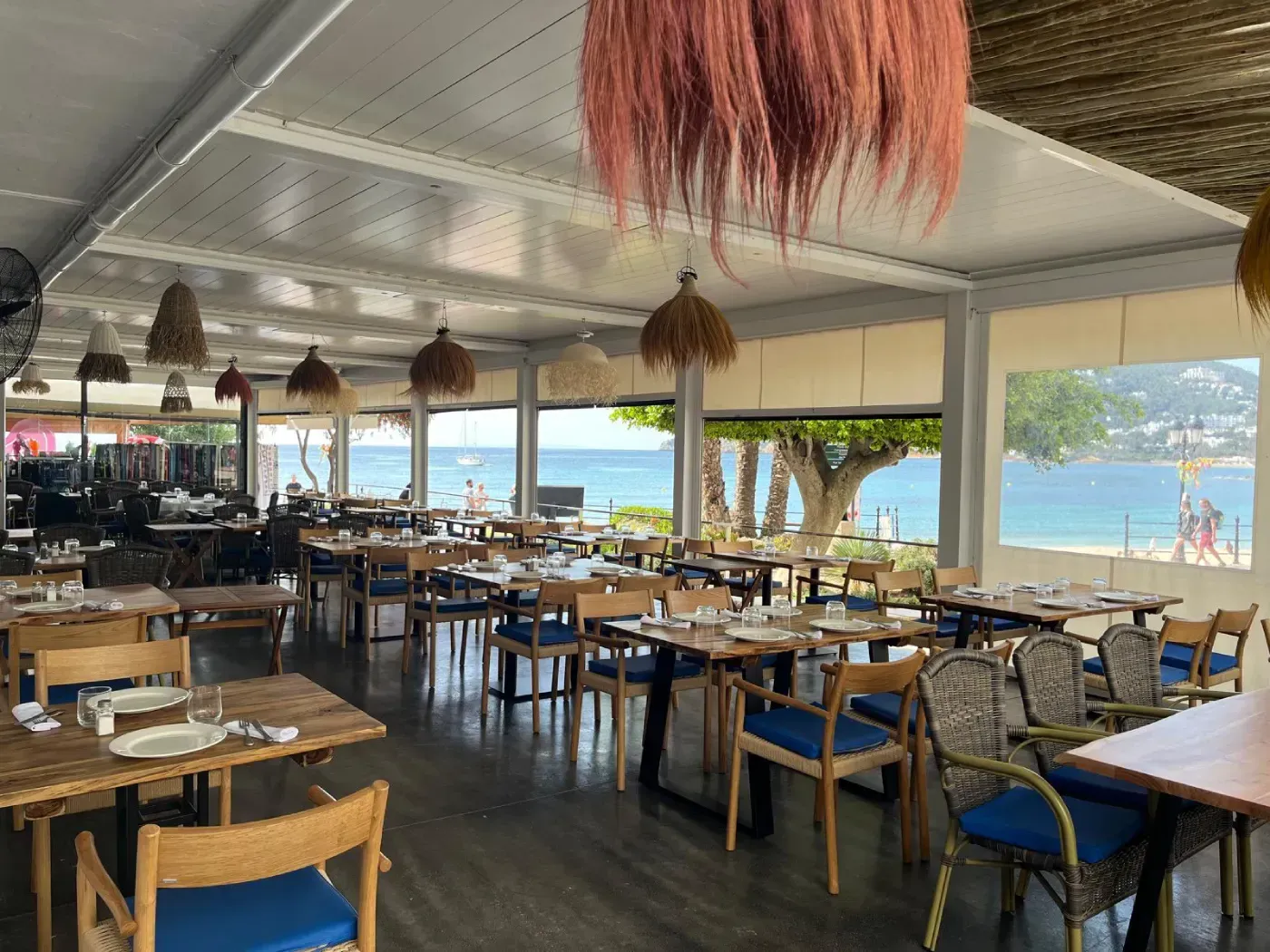 Mesas redondas de comedor dispuestas con platos y sillas de mimbre en un comedor de restaurante vacío.