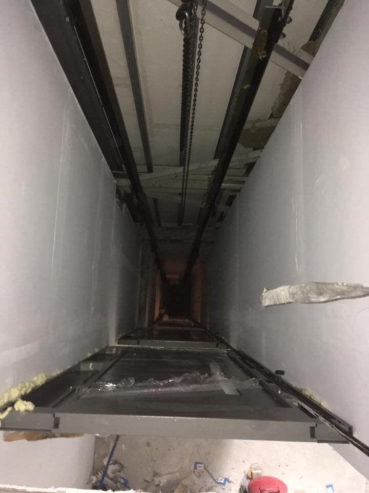 Μέσα σε ένα φρεάτιο ανελκυστήρα, θέα σε μεταλλικές ράγες και ένα άνοιγμα πλατφόρμας ανάμεσα σε λευκούς τοίχους.