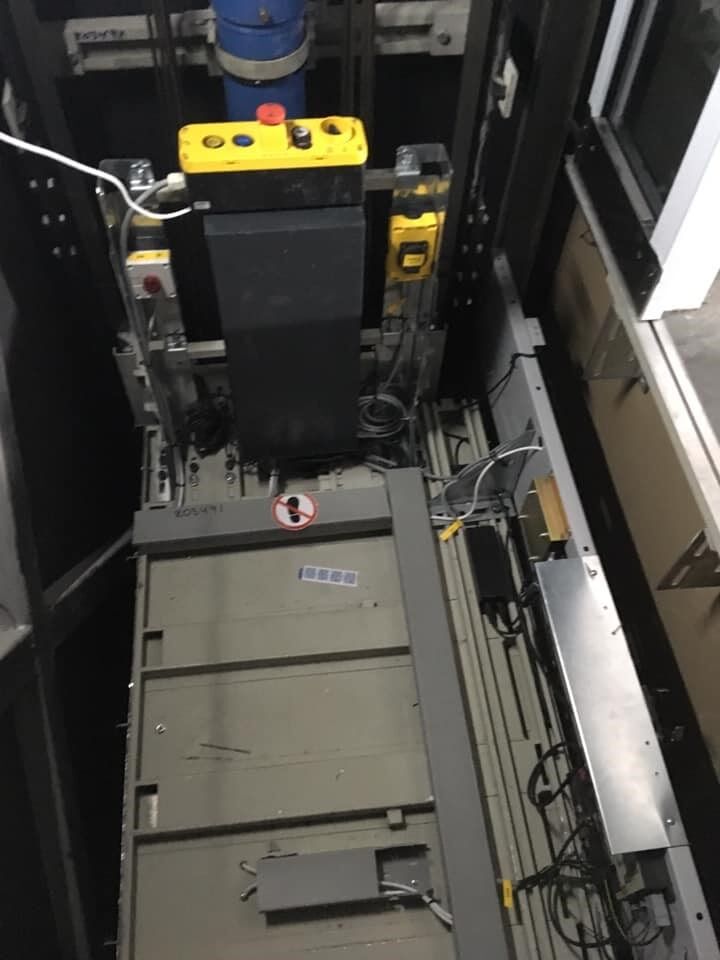 Εσωτερικό φρεατίου ανελκυστήρα με μηχανήματα, καλώδια και γκρίζα πλατφόρμα κάτω από ένα κίτρινο κιβώτιο ελέγχου