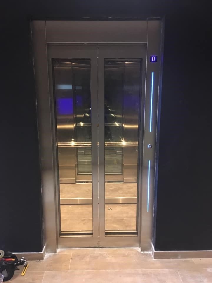 Γυάλινες πόρτες ανελκυστήρα σε έναν σκοτεινό διάδρομο, με ένα μπλε φωτιζόμενο κουμπί κλήσης δίπλα στο πλαίσιο.