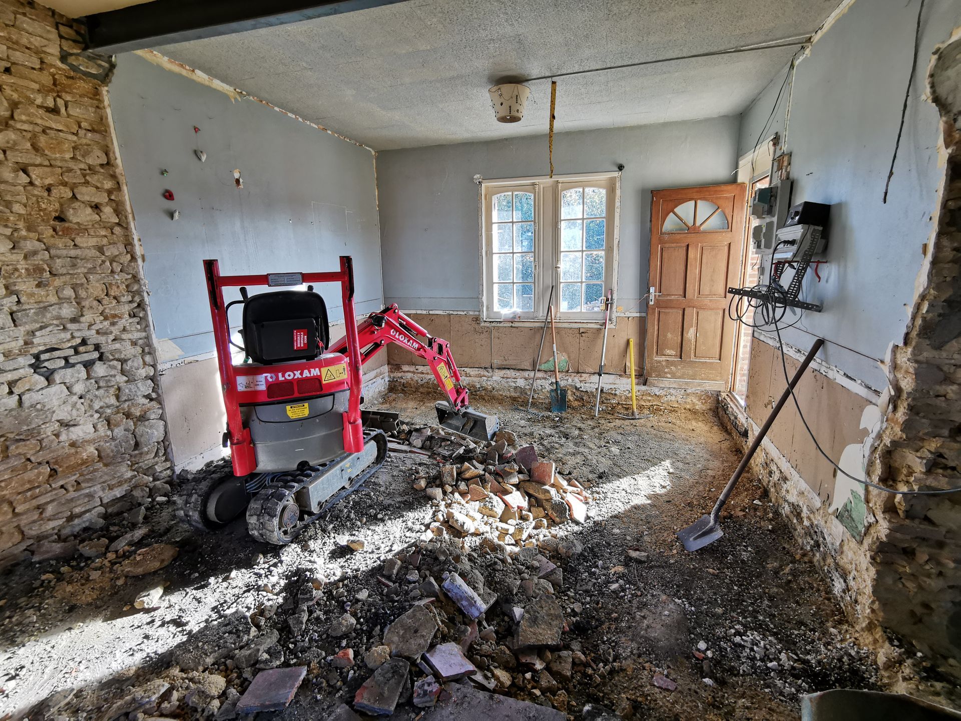 Démolition intérieure à l'aide d'une mini-pelle, gravats au sol, murs, fenêtre et porte mis à nu.