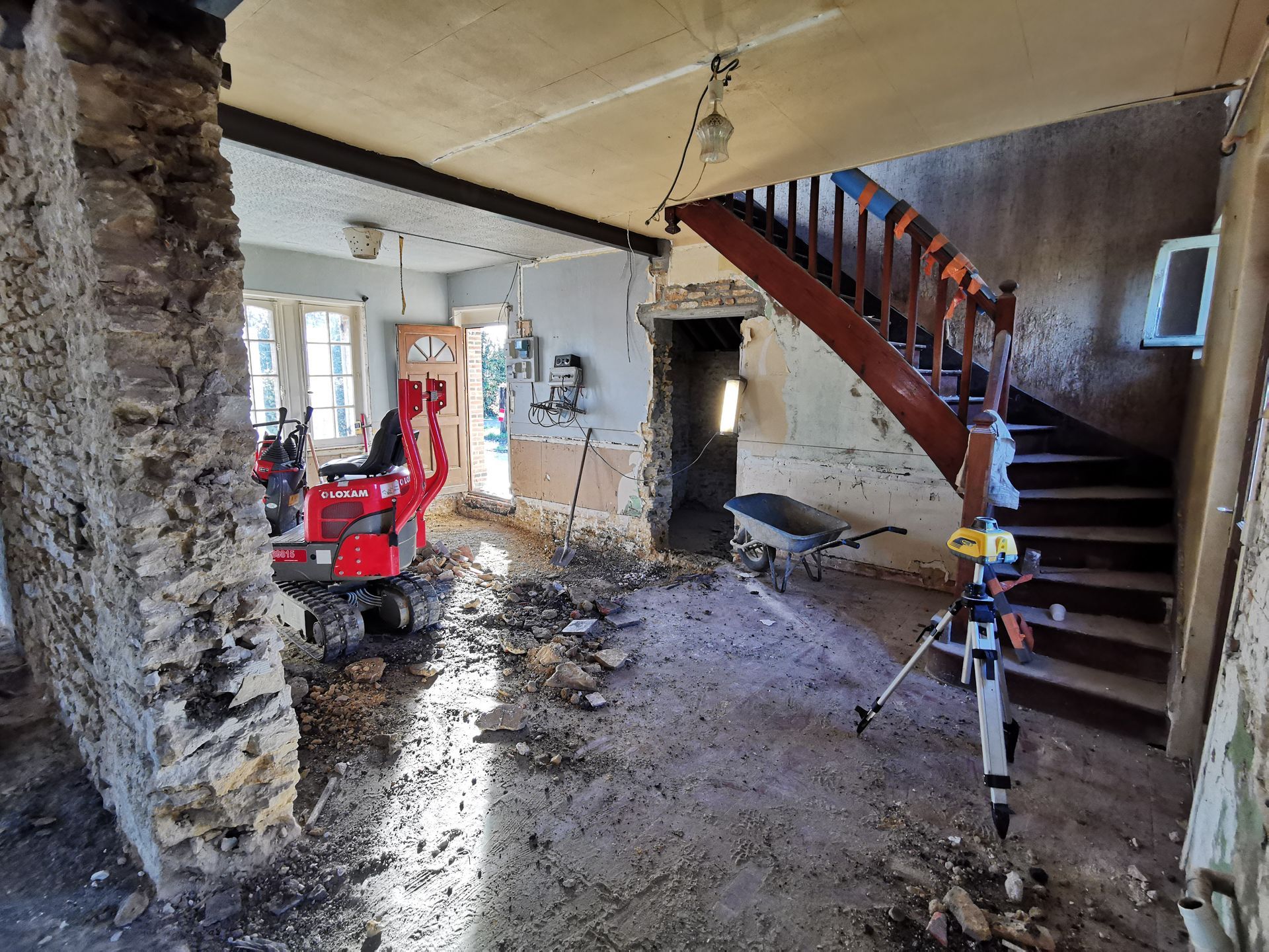 Intérieur en cours de démolition ; mur en pierre apparente, débris au sol, mini-pelle rouge, escalier.