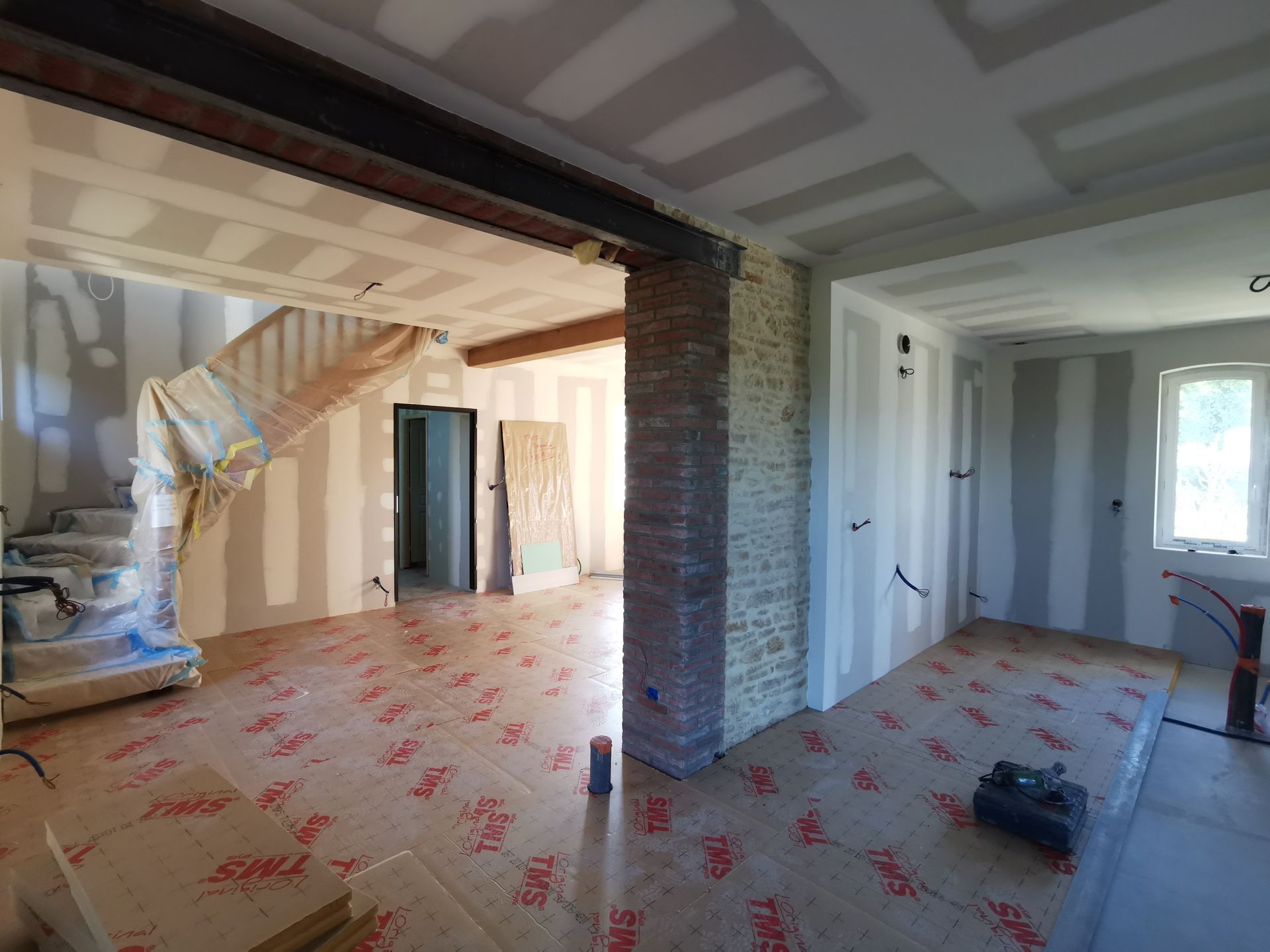 Intérieur d'une maison en construction ; poutres apparentes, colonne en briques, cloisons sèches et revêtement de sol sont visibles.