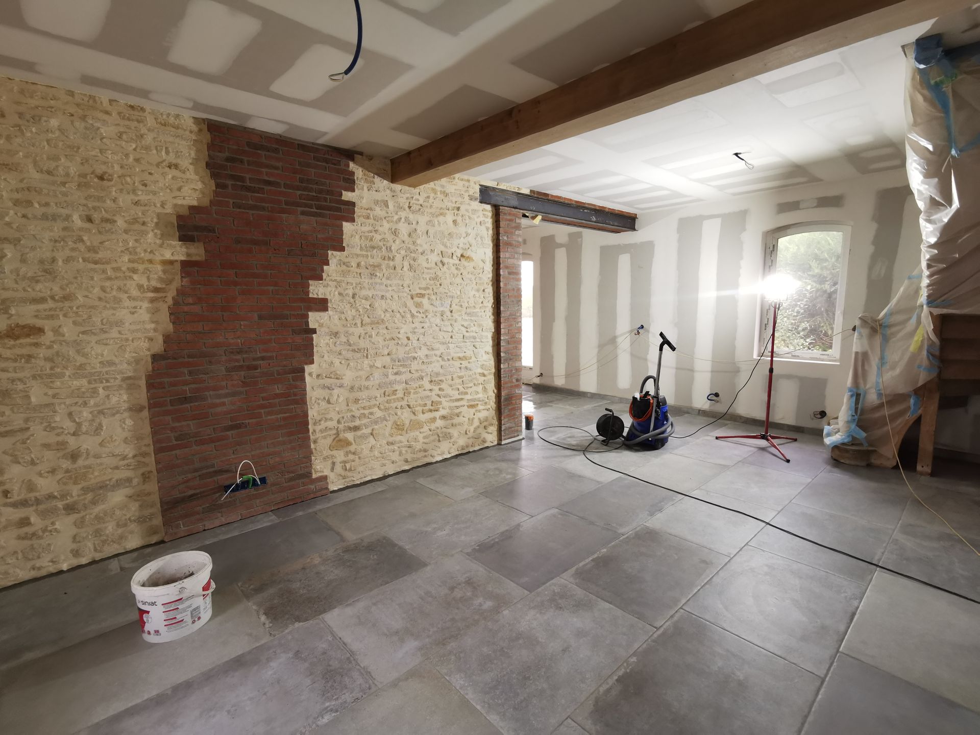 Pièce rénovée : murs en pierre et en brique, sol carrelé gris, poutres apparentes, seau de peinture.