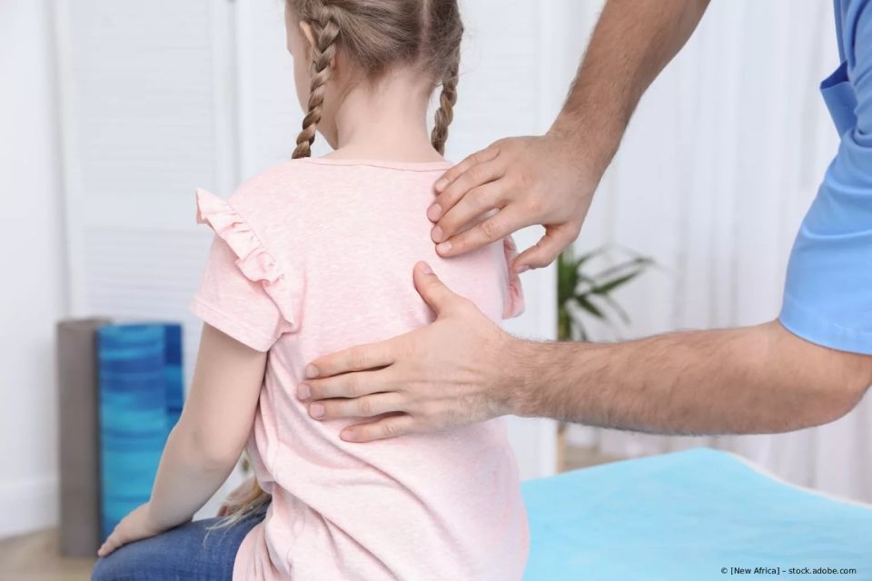 Ein Arzt untersucht den Rücken eines Kindes. Das Kind trägt ein rosa Hemd, der Arzt ein blaues.