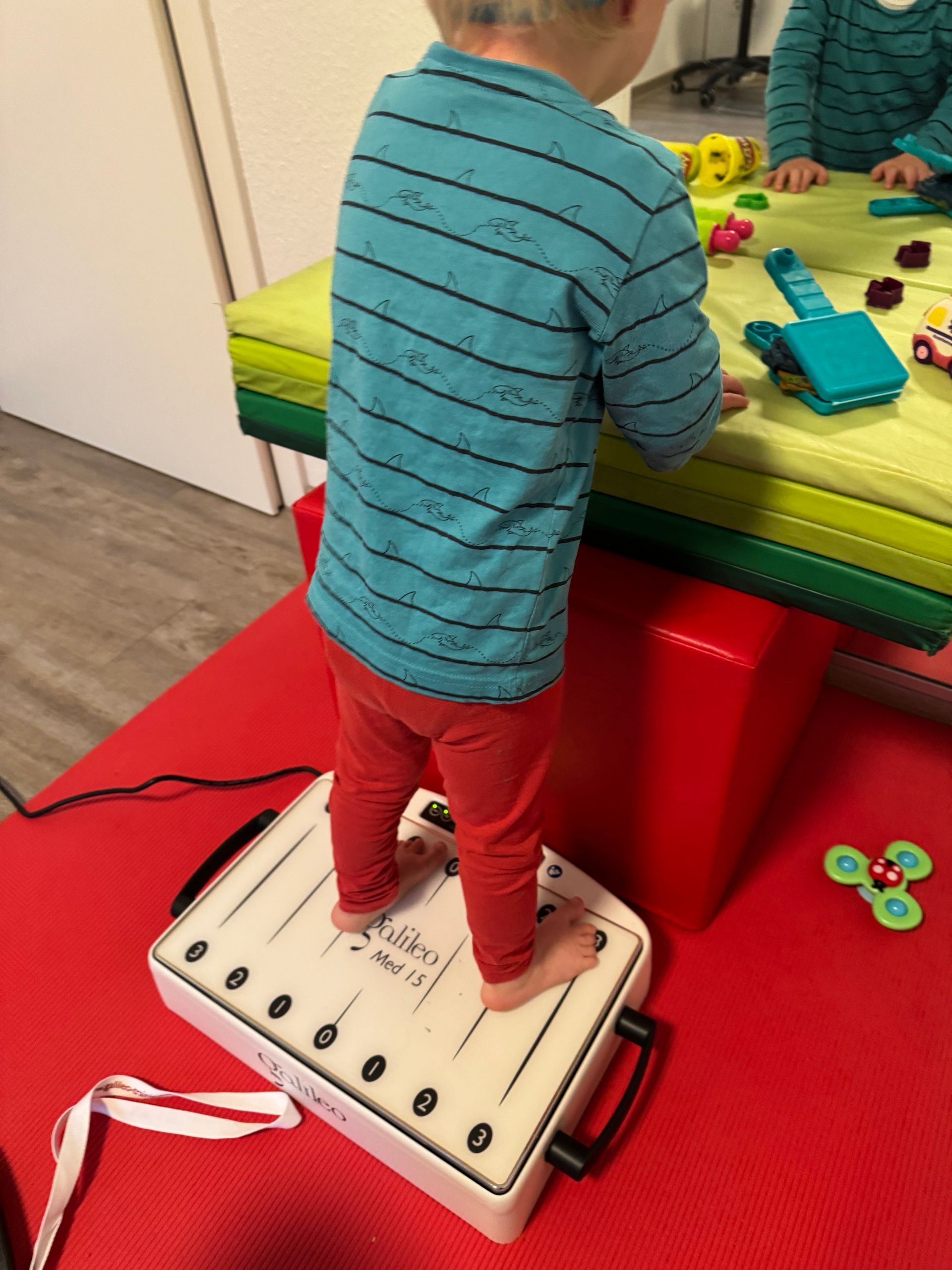 Frau hilft Kleinkind auf Vibrationsplattform in einem Therapieraum.