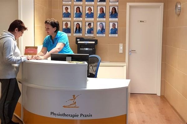 Eine Rezeptionistin betreut einen Patienten an einem orange-weißen Empfangstresen in einer Physiotherapiepraxis.