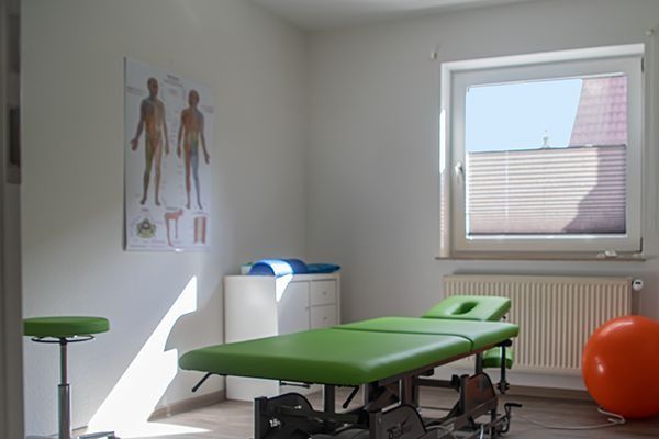 Physiotherapieraum mit Behandlungsliege, anatomischer Tafel, Gymnastikball und Hocker am Fenster.