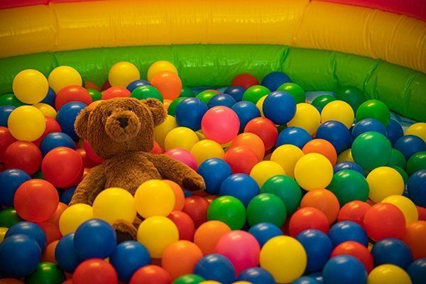 Teddybär in einem bunten Bällebad; gelbe, blaue, rote, grüne und orangefarbene Bälle füllen das Bällebad.