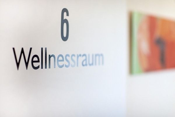 Schild für Raum 6, „Wellnessraum“, an einer weißen Wand; im Hintergrund ein abstraktes Gemälde.