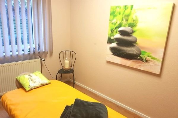Massageraum mit gelber Massageliege, Stuhl mit Kerze und Steinkunstwerk an der Wand.