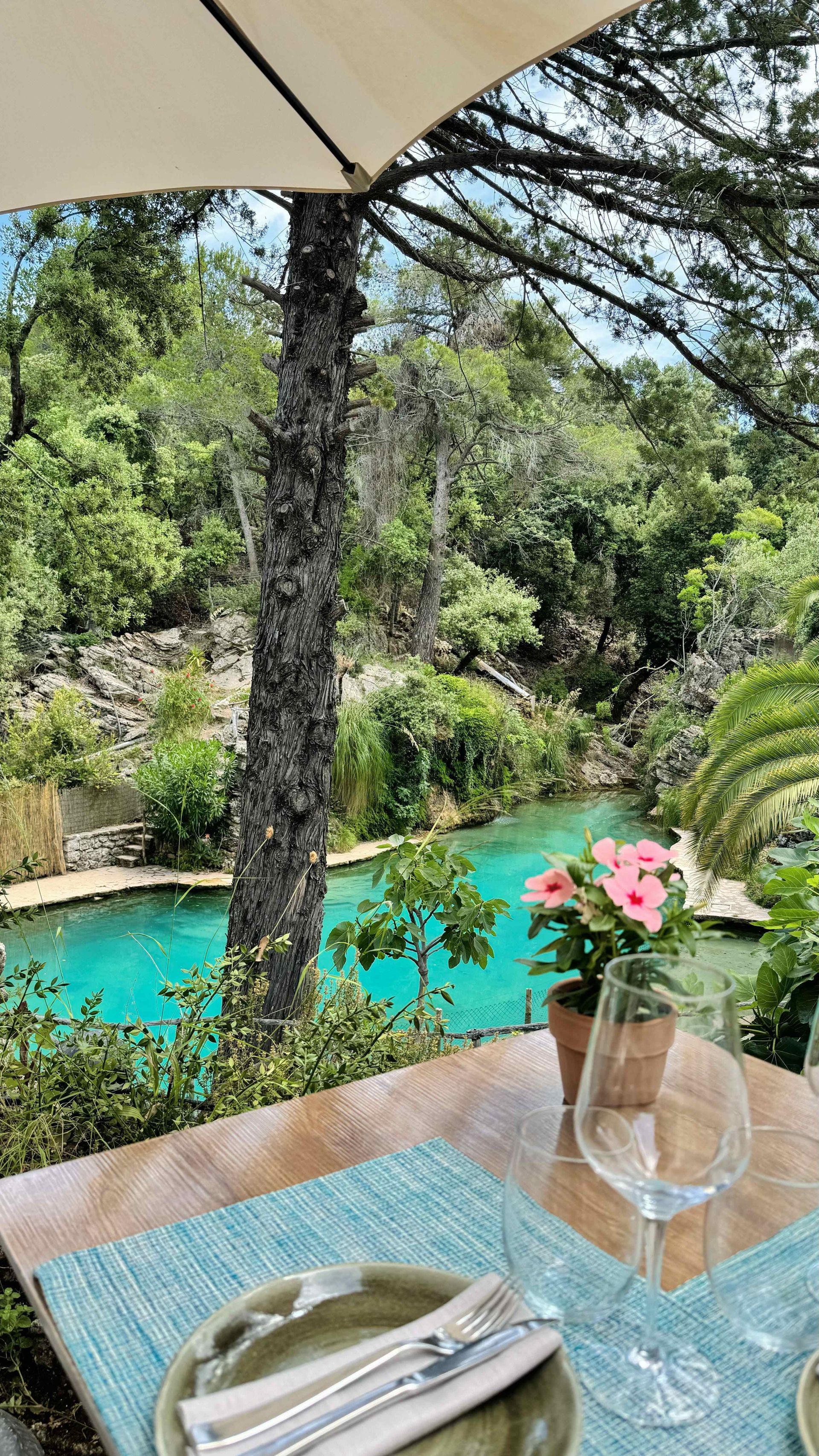 Una mesa con platos, cubiertos, copas de vino y una sombrilla con vistas a una piscina.