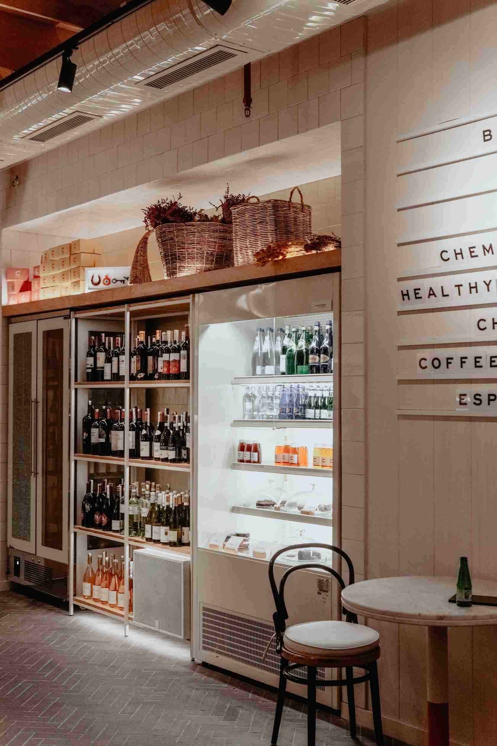 Interior de cafetería con bebidas en vitrina refrigerada, vino y una pequeña mesa con una silla.