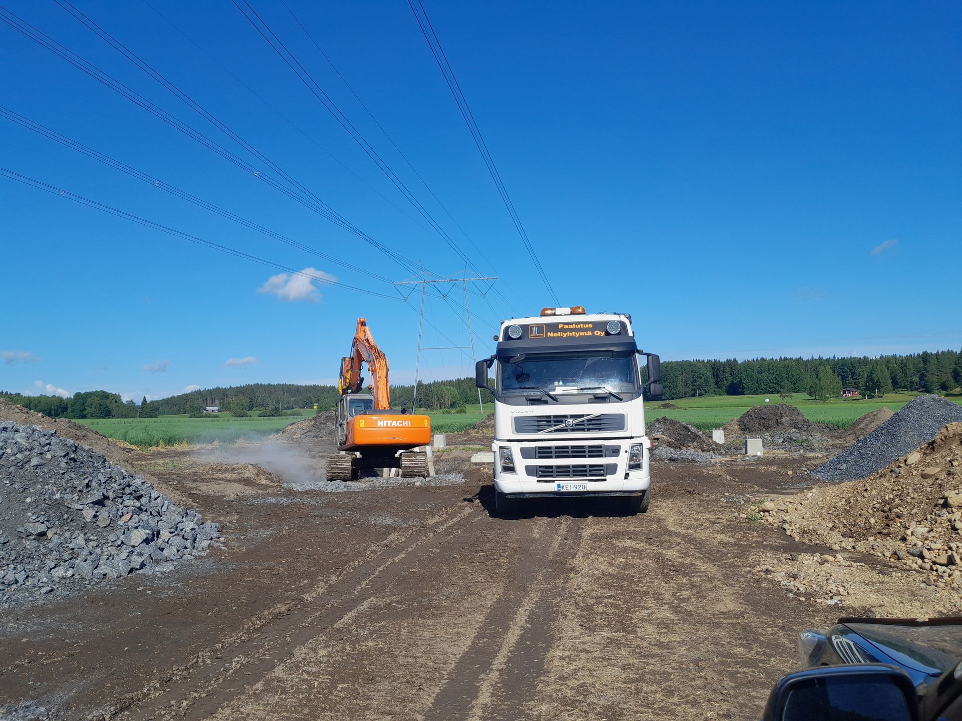 Kaivinkone ja valkoinen kuorma-auto mutaisella rakennustyömaalla kirkkaan sinisen taivaan alla