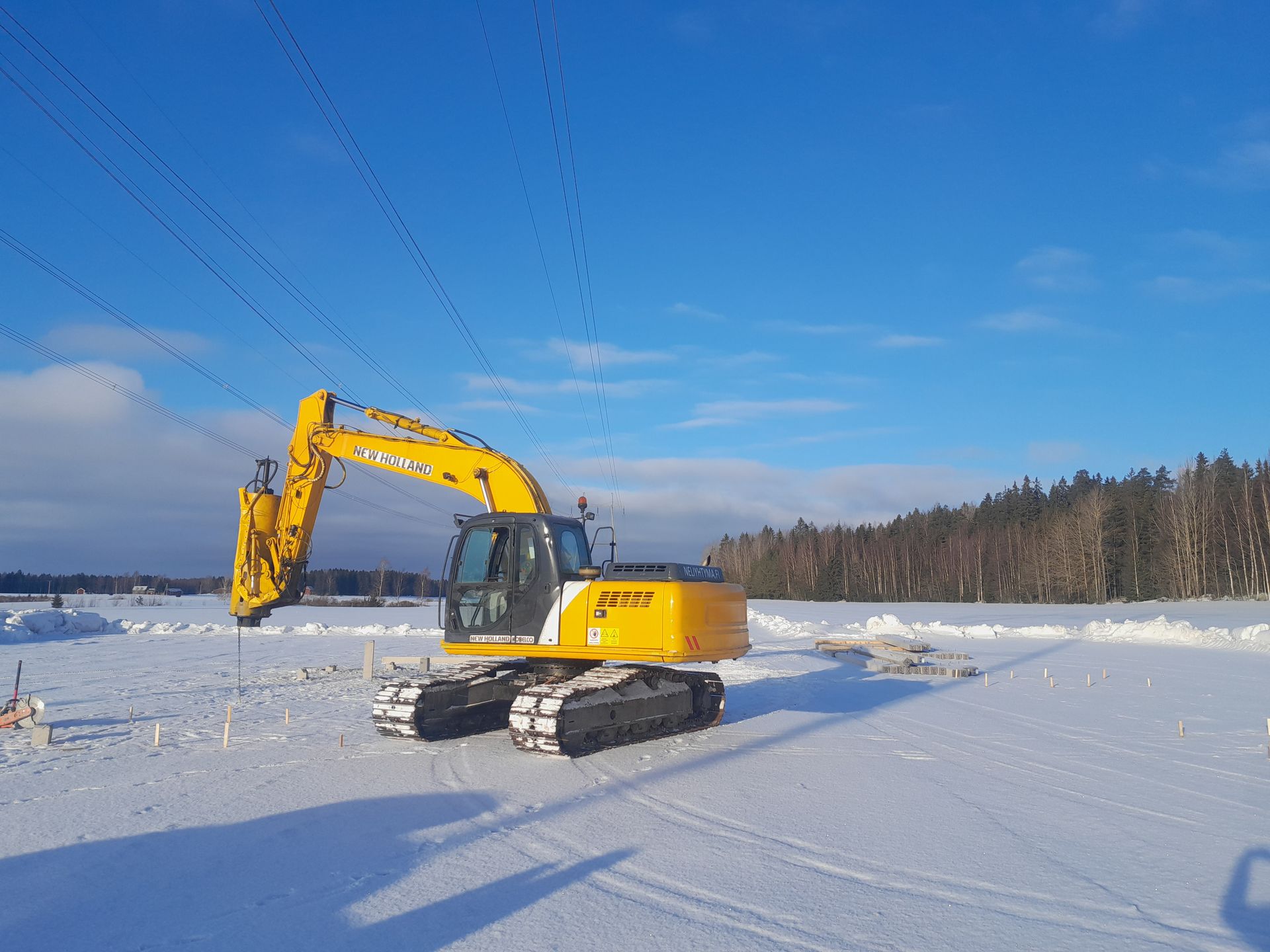 Keltainen kaivinkone lumisella pellolla sinisen taivaan alla, taustalla metsä
