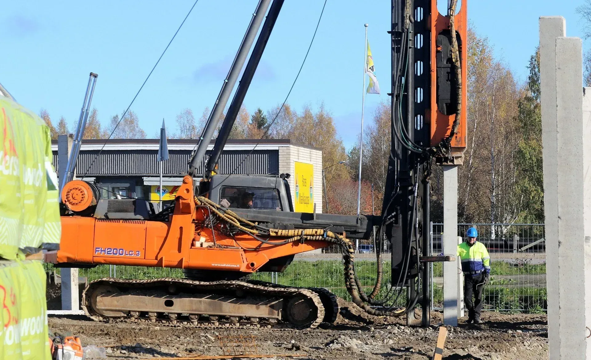 Oranssi porauslautta rakennustyömaalla, ja lähistöllä työntekijä heijastinliiveihin pukeutuneena.