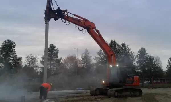 Oranssi kaivinkone porauslaitteella poraa maahan työntekijän vieressä sumuisella rakennustyömaalla