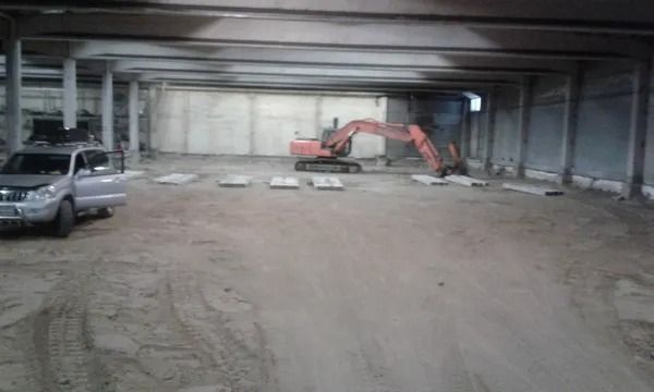 Kaivinkone suuressa keskeneräisessä varastossa pysäköidyn maastoauton vieressä, lattialla rakennusmateriaaleja.