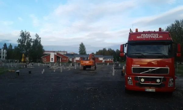Punainen paloauto pysäköitynä tontille pienen rakennuksen lähelle ja oranssi kuorma-auto pilvisen taivaan alla